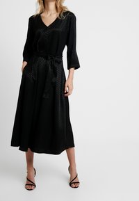 Abito midi nero in tessuto testurizzato con scollo a V, maniche a tre quarti, cintura in vita e tasche laterali. Indossato con tacchi neri con cinturini.