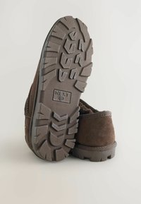 Brune ruskind slip-on sko med en struktureret gummisål, der har et skridsikkert mønster. Sålen er grå og præget med mærkenavnet "NEXT."
