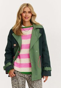 Zelená a černá nepromokavá bunda s velkým límcem, spárovaná s pruhovaným svetrem v růžové a krémové barvě a kalhotami s leopardím potiskem.