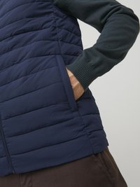 Gilet matelassé marine avec des coutures verticales, une texture horizontale et une poche latérale. Porté par-dessus une chemise à manches longues vert foncé.