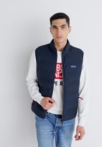 Marinblå puffväst med ståkrage, framdragkedja och sidofickor. Bärs över en vit sweatshirt med röd text. Avslappnad stil.