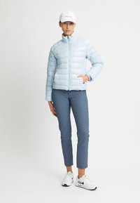 Chaqueta de plumas azul claro con costuras horizontales, cuello alto y cremallera frontal. Combinada con pantalones azules con estampado y zapatillas blancas.
