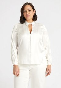 Camicia bianca a maniche lunghe con colletto, strisce verticali e pattina centrale con bottoni. Presenta un'apertura a forma di chiave e una texture morbida.
