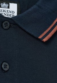 Tamsiai mėlynas polo marškinėliai su ruda juostele ant apykaklės, juodais mygtukais, pažymėtais "Weekend Offender", ir daliniu baltu drabužių etikete su karūnos logotipu.