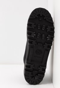 Black rubber shoe sole with deep treads and grooves. Features a circular logo at the bottom center and a textured surface for grip.