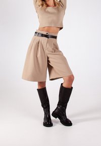 Femme en top court beige et short large plissé avec une ceinture noire, portant des bottes en cuir noires montantes, se tenant contre un fond blanc.