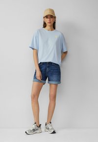 Lichtblauwe t-shirt met een kleine zonafbeelding, gecombineerd met opgerolde denim shorts en zilveren sportschoenen. Een beige pet maakt de look compleet.