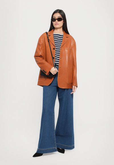 Brown leather blazer, striped shirt, wide-leg blue jeans, and black suede shoes. Black sunglasses and small black bag complete the look.