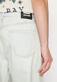Light blue denim jeans with a relaxed fit, featuring a back pocket with frayed detail and a black label reading "DRDENIM" sewn on the waistband.