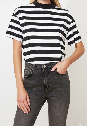 Vrouw die een zwart-wit horizontaal gestreept shirt met korte mouwen draagt, ingestopt in hoge taille donkergrijze spijkerbroek, met één hand in de zak.