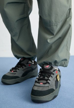 Baskets gris foncé et noires avec des accents rouges portées avec un pantalon vert olive clair sur un sol bleu.