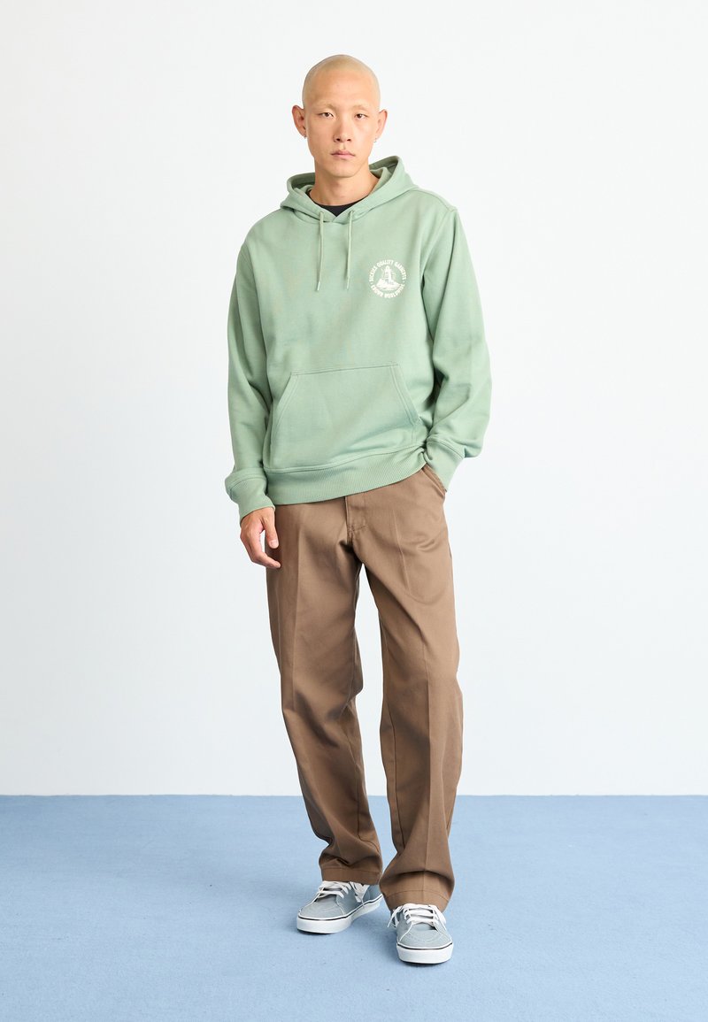 Green hoodie with a front pocket and white logo, paired with brown trousers and gray sneakers. Simple style against a light background.
