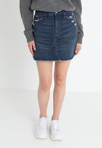 Falda de mezclilla en azul oscuro con dobladillo deshilachado, detalles desgastados y un diseño ajustado. Combinada con zapatillas blancas y un suéter gris.
