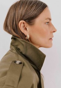 Oliwkowy płaszcz z wysokim kołnierzem, guziki na ramieniu; złote kolczyki o skomplikowanym wzorze; widoczne jasnobrązowe włosy.