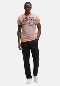 Poloshirt in koraal met zwarte grafische tekst, twee knopen en een kraag. Gecombineerd met zwarte broek en witte sneakers. Neutrale achtergrond.