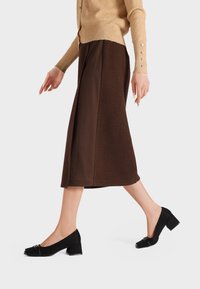 Scarpe con tacco in suede nero e punta quadrata, con una fibbia decorativa. Indossate con una gonna midi marrone e un cardigan beige, mostrate in movimento.