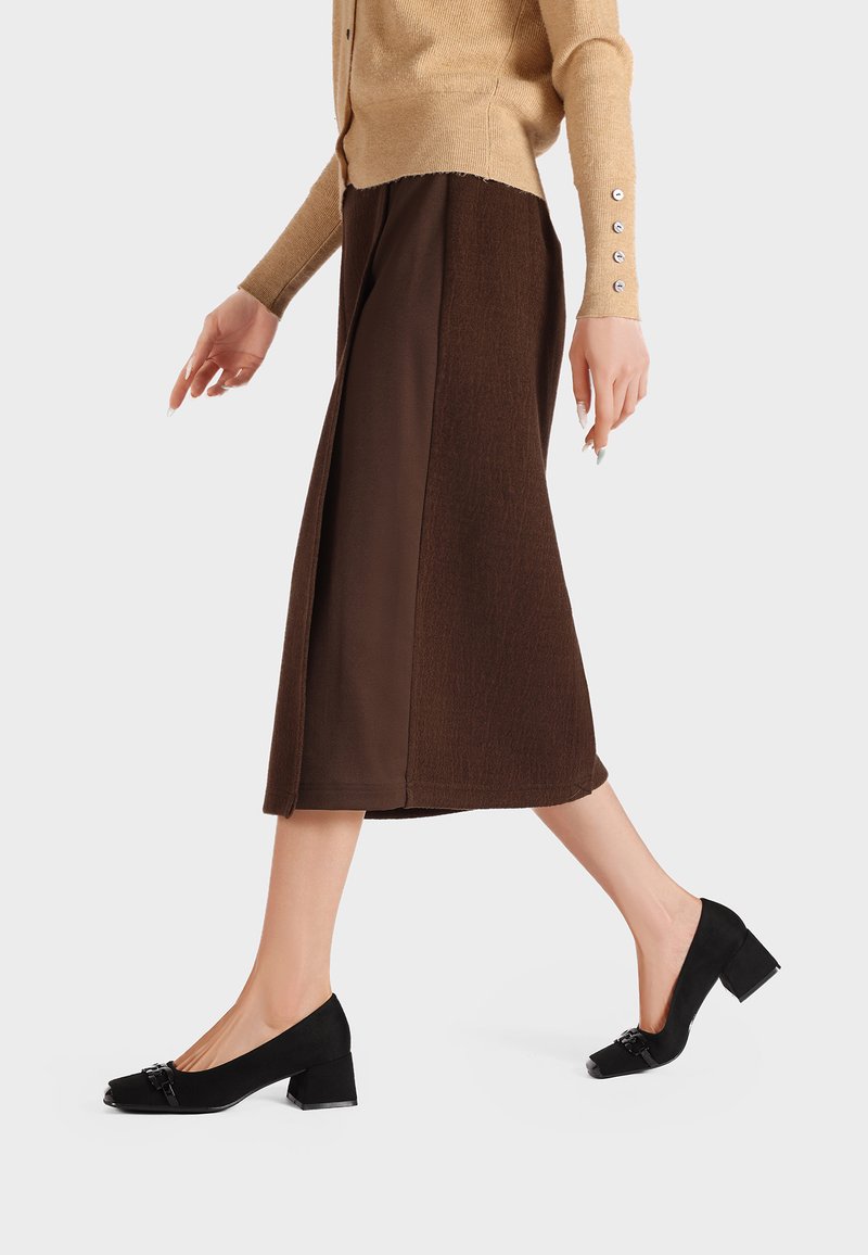 Scarpe con tacco in suede nero e punta quadrata, con una fibbia decorativa. Indossate con una gonna midi marrone e un cardigan beige, mostrate in movimento.