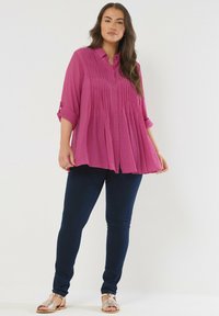 Camisa fucsia de botones con diseño plisado y mangas enrolladas, combinada con jeans pitillo azul oscuro y sandalias metálicas.