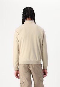 Veste beige légère avec un col montant, des poignets et un ourlet élastiques, vue de dos. Le tissu est lisse avec une texture douce.
