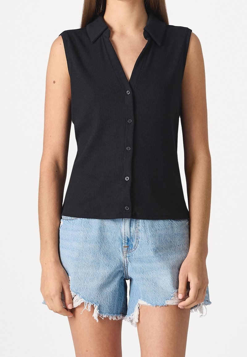 Chemise noire sans manches à boutons avec col, portée avec un short en jean bleu clair effiloché sur une personne, sur un fond blanc uni.