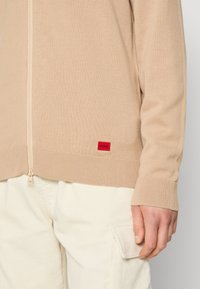 Pull beige zippé avec poignets et ourlet côtelés, arborant un petit logo rouge Hugo près de la taille, porté avec un pantalon de couleur claire.