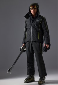 Chaqueta de esquí negra con acentos amarillos, combinada con pantalones negros y botas de esquí. El esquiador sostiene esquís y lleva gafas. Tela texturizada visible.