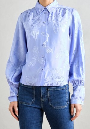Kvinde iført lys lilla blomstret skjorte med knapper, puffede lange ærmer og mørkeblå jeans.