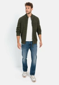 Dunkelgrüner, strukturierter Cardigan mit Stehkragen und Reißverschluss, getragen über einem weißen T-Shirt, kombiniert mit blauen Jeans und hellen Sneakers.