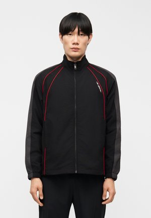 Férfi fekete cipzáras dzsekiben, piros szegéllyel és szürke csíkkal az ujjakon, egyszínű háttér előtt.