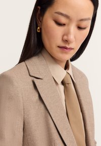 Beige Fischgrät-Blazer mit strukturierten Schultern, kombiniert mit einem hellbraunen Hemd und passender Krawatte; goldene Creolen als Akzent.