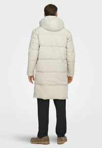 Lichtbeige pufferjas met een capuchon, met een doorgestikt ontwerp, middellange silhouet en elastische manchetten, gecombineerd met zwarte broek en bruine schoenen.