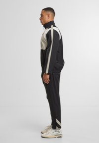 Tuta nera e beige con colletto alto, caratterizzata da un design a blocchi di colore diagonali e polsini elasticizzati, abbinata a sneaker di colore chiaro.