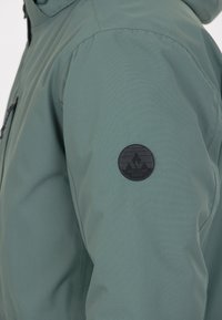 Grön jacka med slät, matt textil och en rund svart logotyp på vänster ärm. Dragkedjefickor synliga fram.