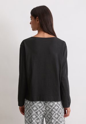 Vrouw met lang donker haar draagt een eenvoudige zwarte top met lange mouwen en een witte broek met zwarte bloemen- en ruitpatronen, met de rug naar de camera gekeerd.