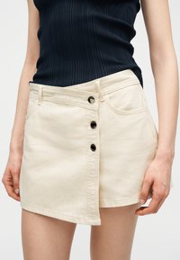 Femme portant une mini-jupe asymétrique crème à boutons sur le devant et un haut noir sans manches en maille côtelée, les mains détendues le long du corps.