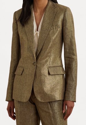 Frau trägt einen maßgeschneiderten Metallic-Gold-Blazer mit Pattentaschen und passenden Hosen über einer weißen Bluse mit Schleifenkragen.
