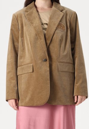 Mujer con una chaqueta de pana marrón claro con solapas con muesca, cierre frontal con botones, bolsillos con solapa y una falda rosa.