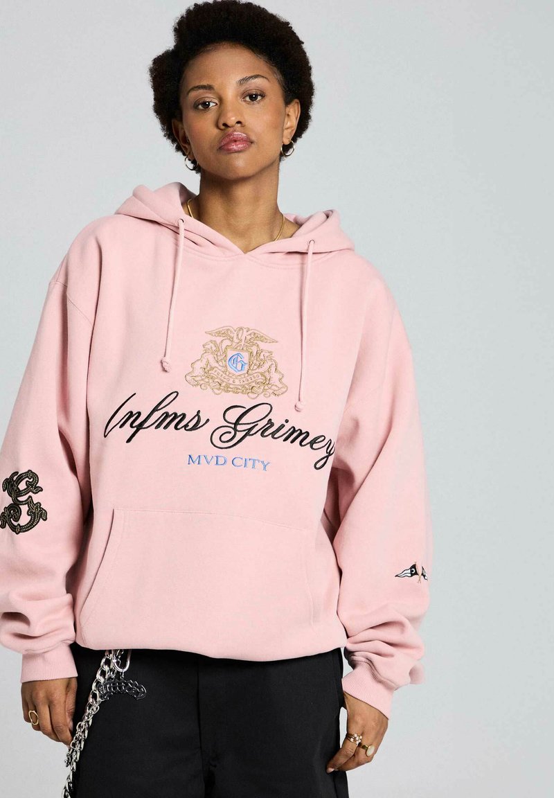 Joven con sudadera con capucha rosa con texto y diseños "Infms Grimey MVD City", combinada con pantalones negros y accesorio de cadena.