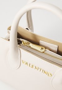 Bolso de cuero crema con dos asas, herrajes dorados, cierre de cremallera y logo en relieve "VALENTINO" en la parte frontal, que muestra una textura suave.