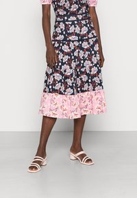 Saia tiered floral com fundo azul-marinho e bainha rosa, apresentando padrões florais brancos e vermelhos. Combinada com sandálias de salto bloco rosa claro.