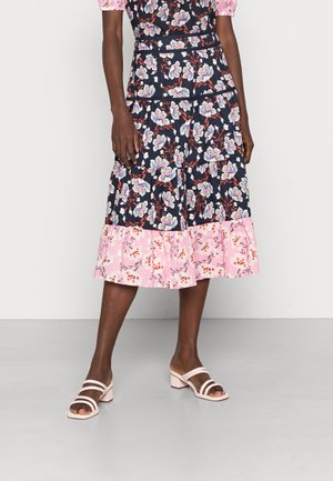 Saia tiered floral com fundo azul-marinho e bainha rosa, apresentando padrões florais brancos e vermelhos. Combinada com sandálias de salto bloco rosa claro.