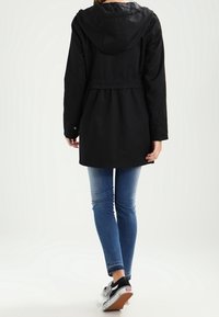 Svart, midjelång jacka med huva, elastisk midja och knäppta manschetter. Bärs över blå jeans och svarta sneakers, med en enkel design.