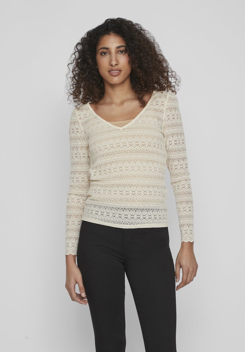 Blonde bluse med lange ermer i kremhvitt med V-hals, med blomster mønster og en tettsittende design. Brukt med svarte bukser.