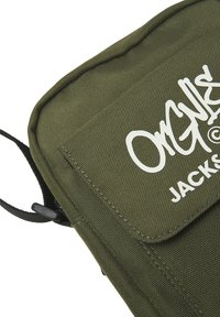 Bolso de hombro de lona en verde oliva. Cuenta con un cierre de solapa, correa negra y logo impreso en blanco. Superficie texturizada con costuras visibles.
