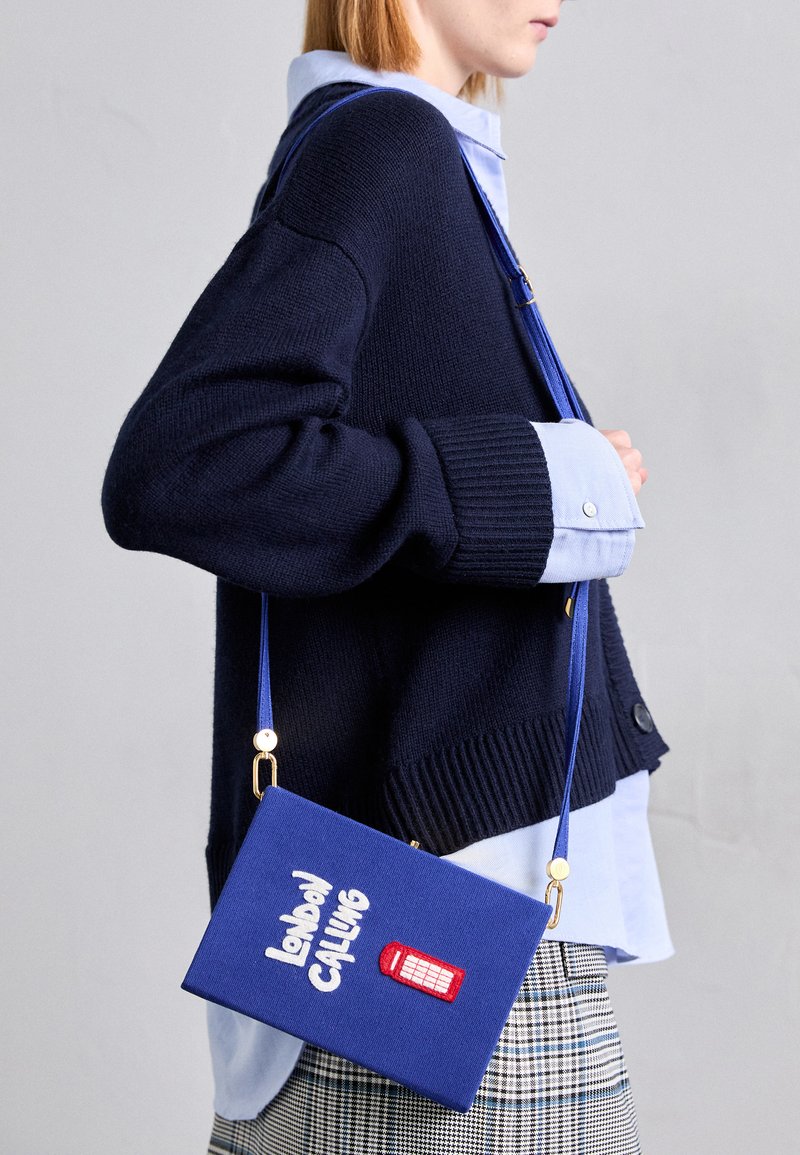 Navy knit sweater, light blue collared shirt underneath. Blue handbag with "London Calling" text and red phone booth design. Plaid pants.