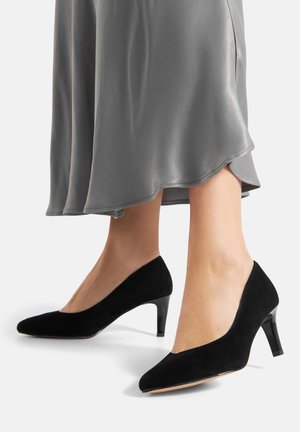 Czarne zamszowe szpilki o wąskim nosku i eleganckim wzornictwie; błyszczący czarny obcas podkreśla sylwetkę buta.