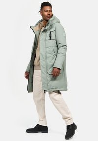Parka de culoare verde deschis, cu o căptușeală moale de culoare bej, buzunare mari în față și glugă, asortată cu pantaloni de culoare crem și pantofi negri.