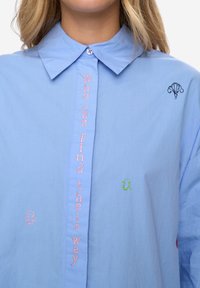 Camicia blu chiaro con colletto appuntito. Presenta ricami rosa e verdi con testo e simboli. Tessuto morbido, vestibilità comoda.