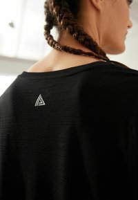 Juodas sportinis marškinėlis su lygiu paviršiumi, ant viršutinės nugaros dalies esantis baltas trikampio formos logotipas. Dizainas apima plačią iškirptę.
