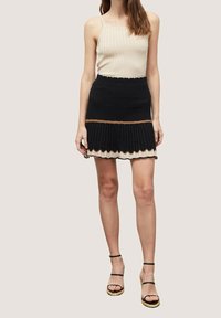 Beige rippad tanktop i kombination med en svart stickad kjol med en vågig nederkant, brun rand och vita vågiga detaljer. Svarta sandaletter med remmar.